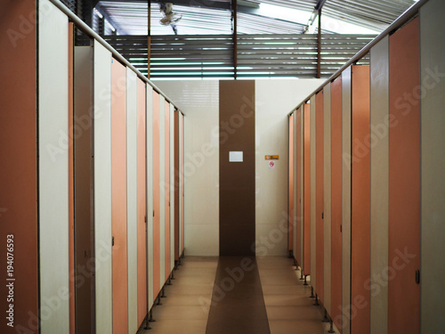 Interior of Thai public restroom in rural gas station, with white wall, many brown pink doors, and wet ceramic floor as center trail walkway with toilet on both side, high ceiling, sunlight