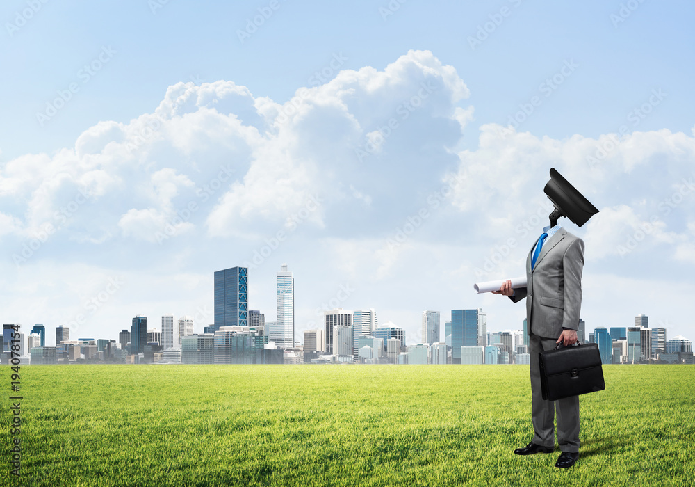Camera headed man standing on green grass against modern cityscape Camera headed man standing on green grass against modern cityscape