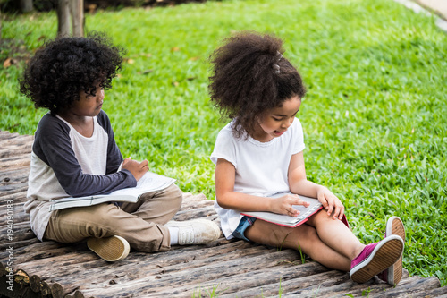 Little boy and girl reading book while sitting on ground in a park.