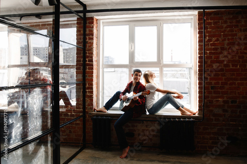 stylish young couple having fun in cozy bedroom in style loft,  feeling happy being together. The man plays the guitar and sings the song for the woman, sitting on a window sill. © Мария Балчугова