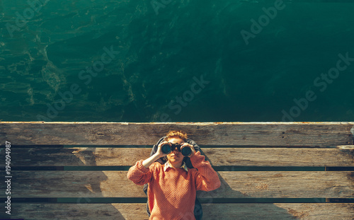 Young Beautiful Girl Lies On A Pier Near The Sea And Looks Through Binoculars On The Sky. Travel Search Journey Concept