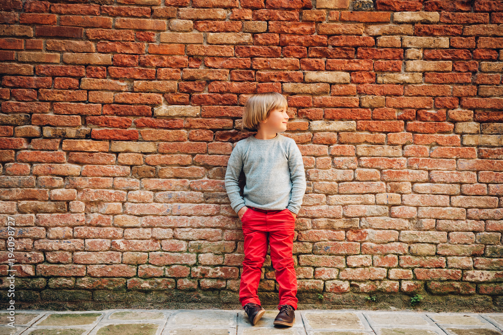 Outdoor portrait of handsome little boy wearing red chinos and light blue pullover, stylish kid posing against vintage brick wall. Fashion for small children Outdoor portrait of handsome little boy wearing red chinos and light blue pullover, stylish kid posing against vintage brick wall. Fashion for small children