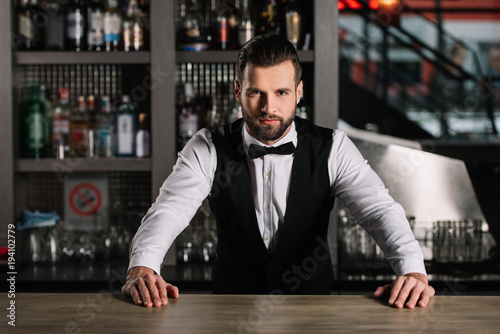 handsome bartender leaning on bar counter and looking at camera photo