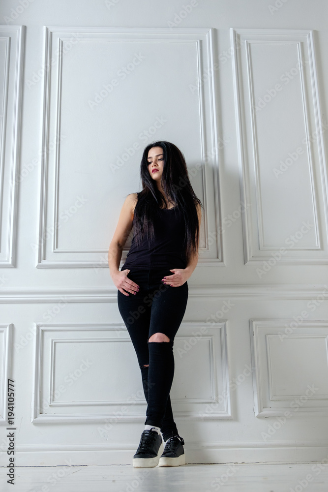 brunette caucasian girl in black casual style clothes standing near wall in white interior brunette caucasian girl in black casual style clothes standing near wall in white interior