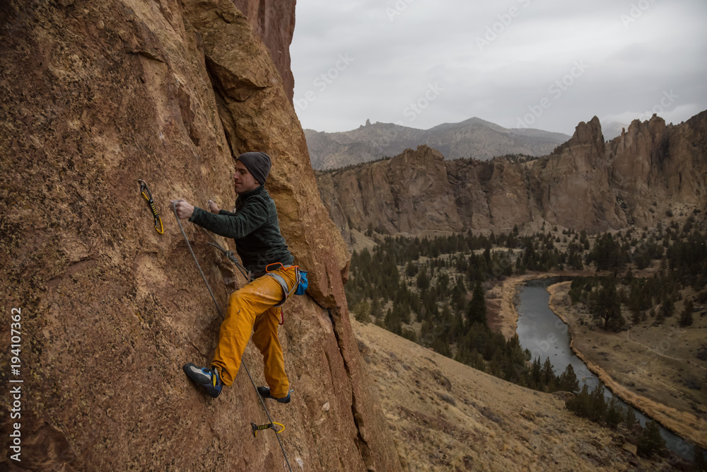 Adventurous man is rock climbing on the side of a steep cliff during a cloudy winter evening. Taken in Smith Rock, Oregon, North America. Adventurous man is rock climbing on the side of a steep cliff during a cloudy winter evening. Taken in Smith Rock, Oregon, North America.