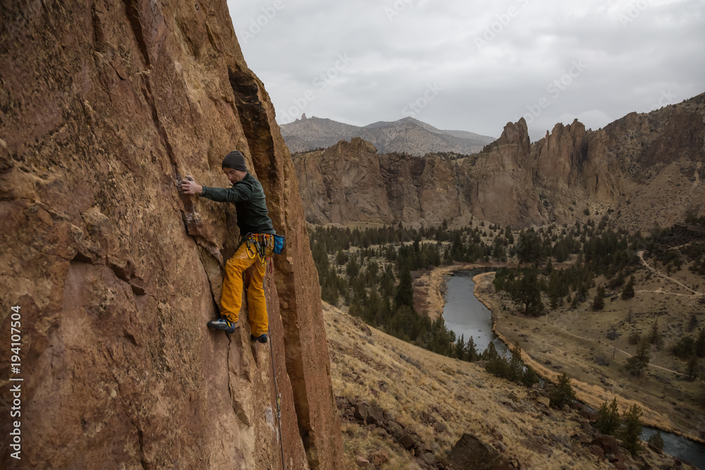 Adventurous man is rock climbing on the side of a steep cliff during a cloudy winter evening. Taken in Smith Rock, Oregon, North America.