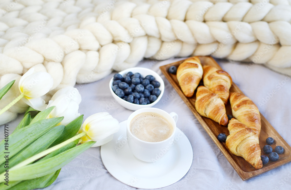 Cup with cappuccino and croissants, berries, white pastel giant knit blanket, bedroom, flowers tulips, spring, woman day, morning concept 