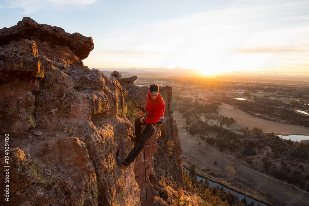 Obraz premium Adventurous man is rappeling down a cliff during a bright and vibrant sunny sunset. Taken in Smith Rock, Oregon, North America.