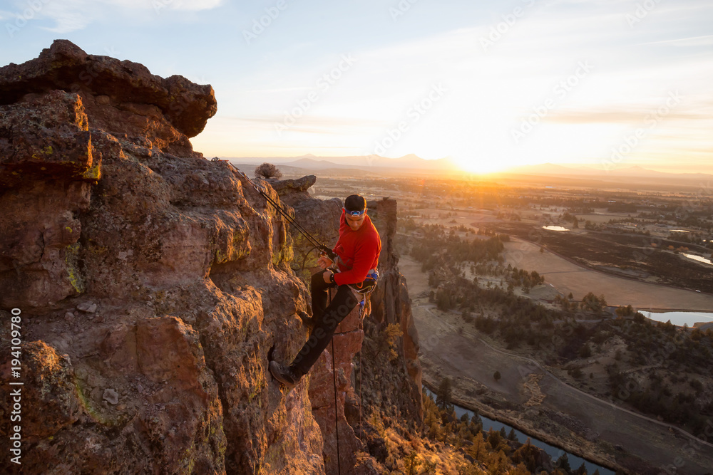 Adventurous man is rappeling down a cliff during a bright and vibrant sunny sunset. Taken in Smith Rock, Oregon, North America.