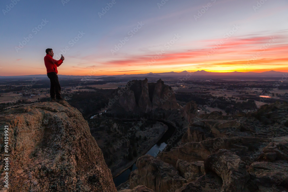 Man standing on top of a mountain is enjoying a beautiful landscape during a colorful and vibrant sunset. Taken at Smith Rock, Oregon, North America.