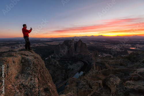 Man standing on top of a mountain is enjoying a beautiful landscape during a colorful and vibrant sunset. Taken at Smith Rock, Oregon, North America.