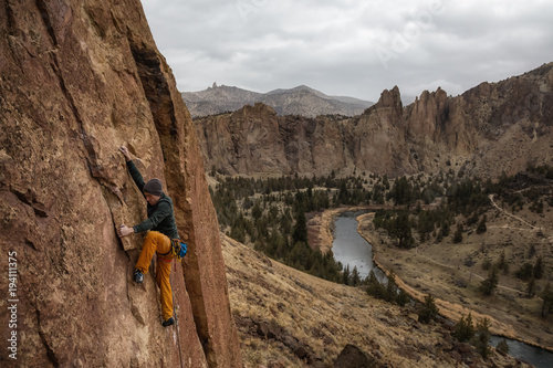 Canvas-taulu Adventurous man is rock climbing on the side of a steep cliff during a cloudy winter evening