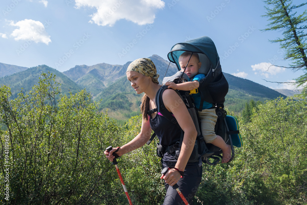 Obraz premium Woman Hiker trekking in mountains with child in backpack . Mother with baby boy travelling in summer sunny day.