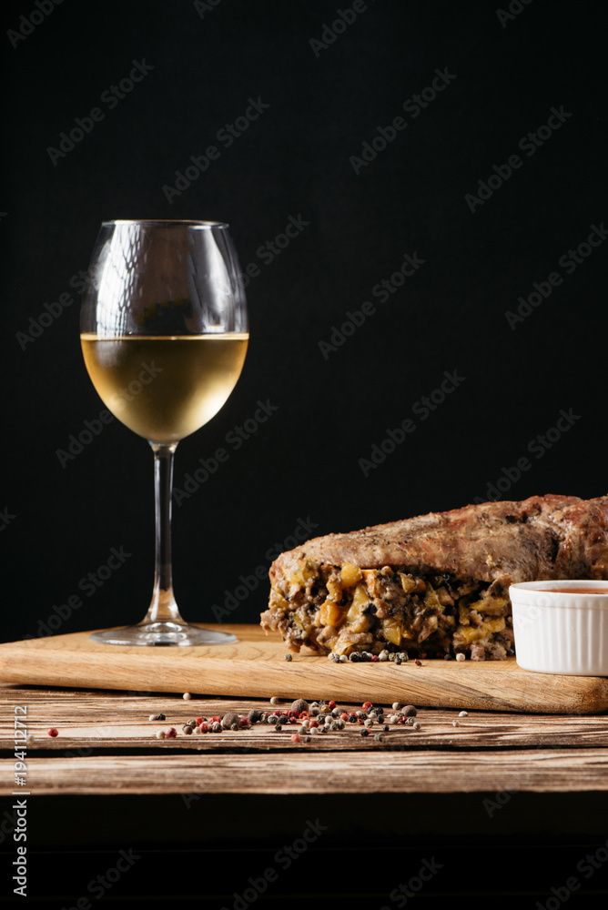 Pumpkin muffin and baked meat roll with filling, along with a sauce on a wooden plate and a glass of white wine on a black background