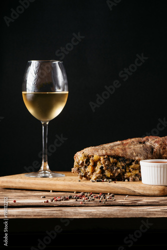Pumpkin muffin and baked meat roll with filling, along with a sauce on a wooden plate and a glass of white wine on a black background