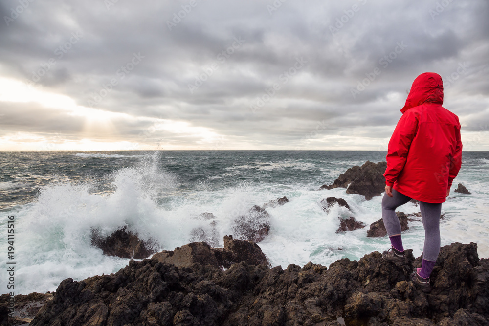 Woman in a bright red jacket is watching waves crash on a rocky Pacific Ocean Coast. Taken in Ucluelet, Vancouver Island, BC, Canada.