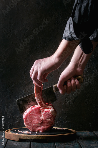 Man's hands cutting raw uncooked black angus beef tomahawk steak on bone by vintage butcher cleaver on round wooden slate cutting board over dark wooden plank table. Rustic style