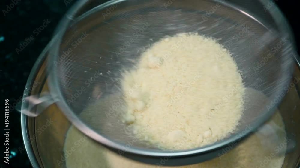 The wheaten flour is being sifted through the big round sieve on the kitchen. The steel bowl is under the strainer which is being shaken and the meal is falling down. The nubbles of powder are jumping The wheaten flour is being sifted through the big round sieve on the kitchen. The steel bowl is under the strainer which is being shaken and the meal is falling down. The nubbles of powder are jumping