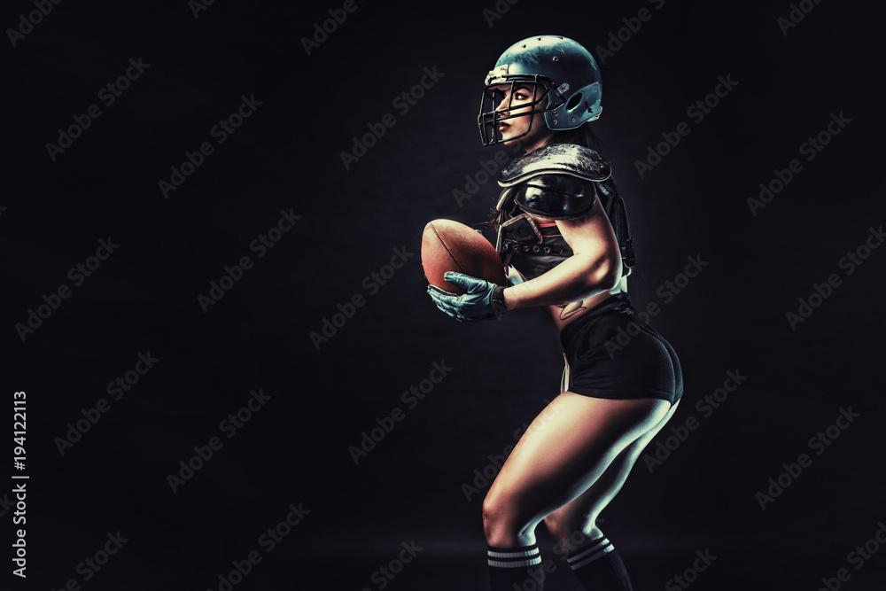 Sportive serious woman in helmet of rugby player holding ball in stuio on dark background.