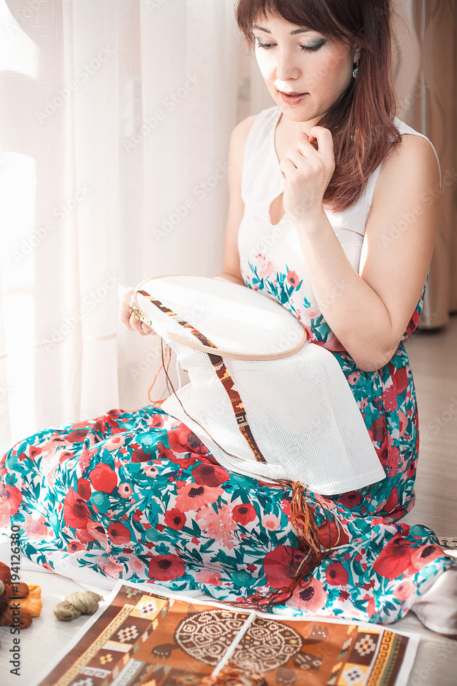 Young beautiful woman is engaged in embroidering a dagger on the embroidery frame in a colored dress at the window in the sunlight. A great picture for a hobby Young beautiful woman is engaged in embroidering a dagger on the embroidery frame in a colored dress at the window in the sunlight. A great picture for a hobby