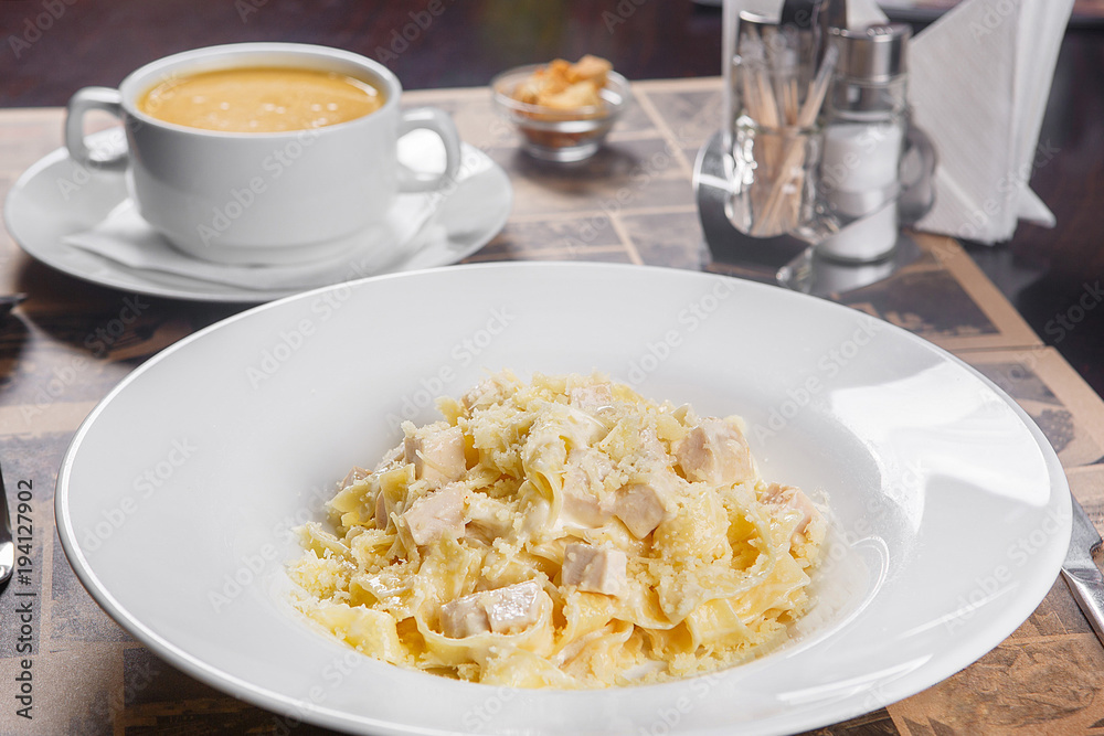 Pasta, noodles with chicken, turkey, decorated with cheese. Serving a meal in a cafe, restaurant. Side view. In the background, soup and crackers, croutons and cutlery Pasta, noodles with chicken, turkey, decorated with cheese. Serving a meal in a cafe, restaurant. Side view. In the background, soup and crackers, croutons and cutlery