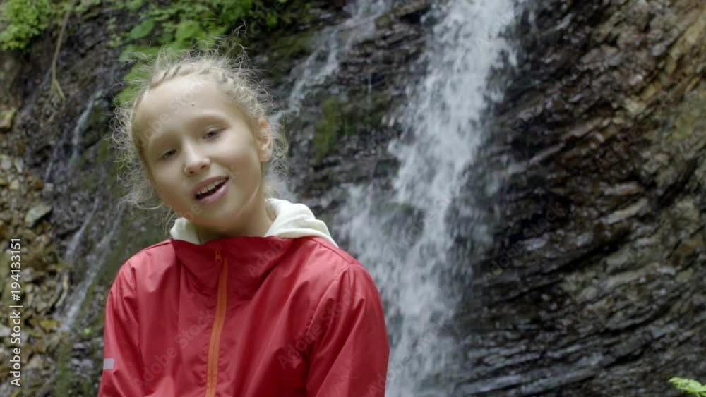 Calm little girl looking at camera, mountain waterfall in forest at background Calm little girl looking at camera, mountain waterfall in forest at background