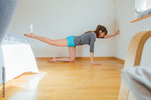 Full length portrait of sporty young woman working out at home in bedroom, doing yoga or pilates exercise on floor. Bird-dog or kneeling opposite arm and leg extension (chakravakasana). Side view photo