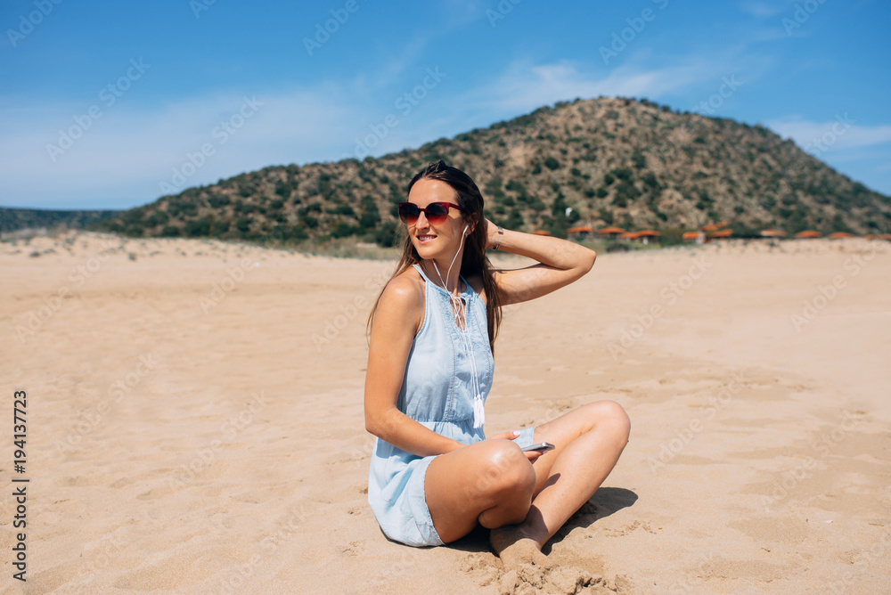 Brunette in sky blue dress sits and looks away in lotus pose on the sand and listens music in the earphones. Green mountain on background Brunette in sky blue dress sits and looks away in lotus pose on the sand and listens music in the earphones. Green mountain on background