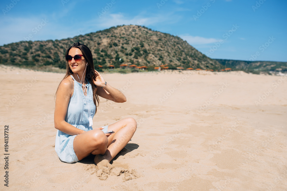 Brunette in sky blue dress sits and looks away in lotus pose on the sand and listens music in the earphones. Green mountain on background