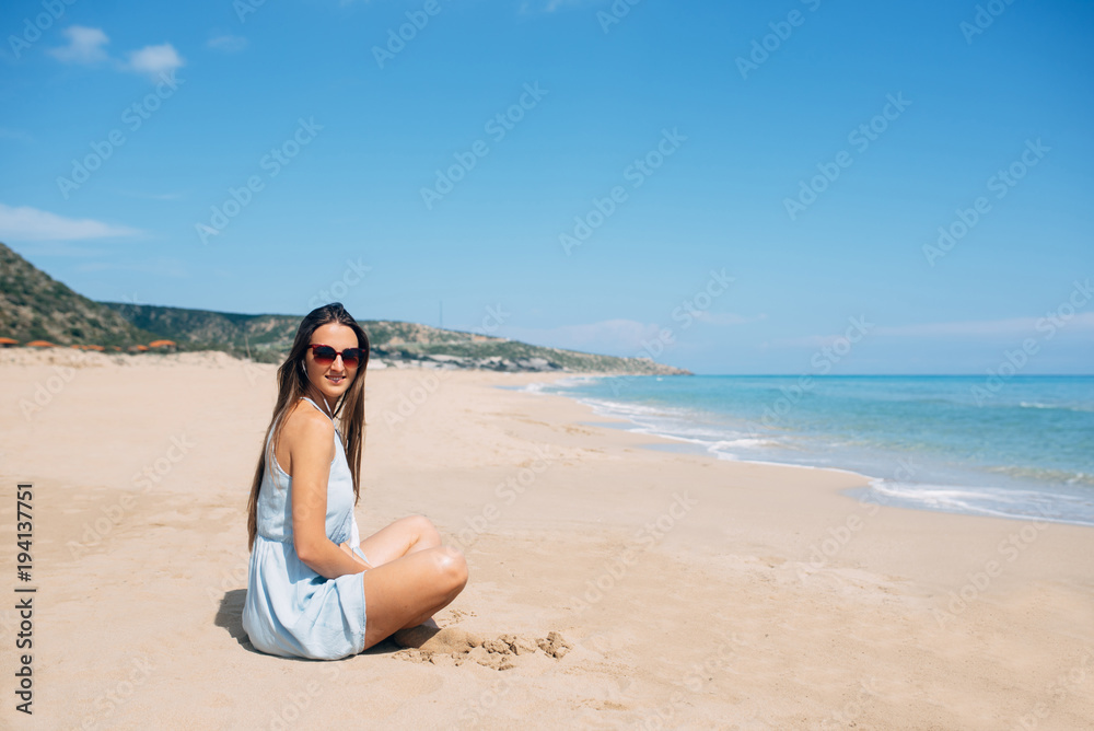 Beautiful young smiling girl sits on the sand and looks in camera. Long beach and sea view on background Beautiful young smiling girl sits on the sand and looks in camera. Long beach and sea view on background