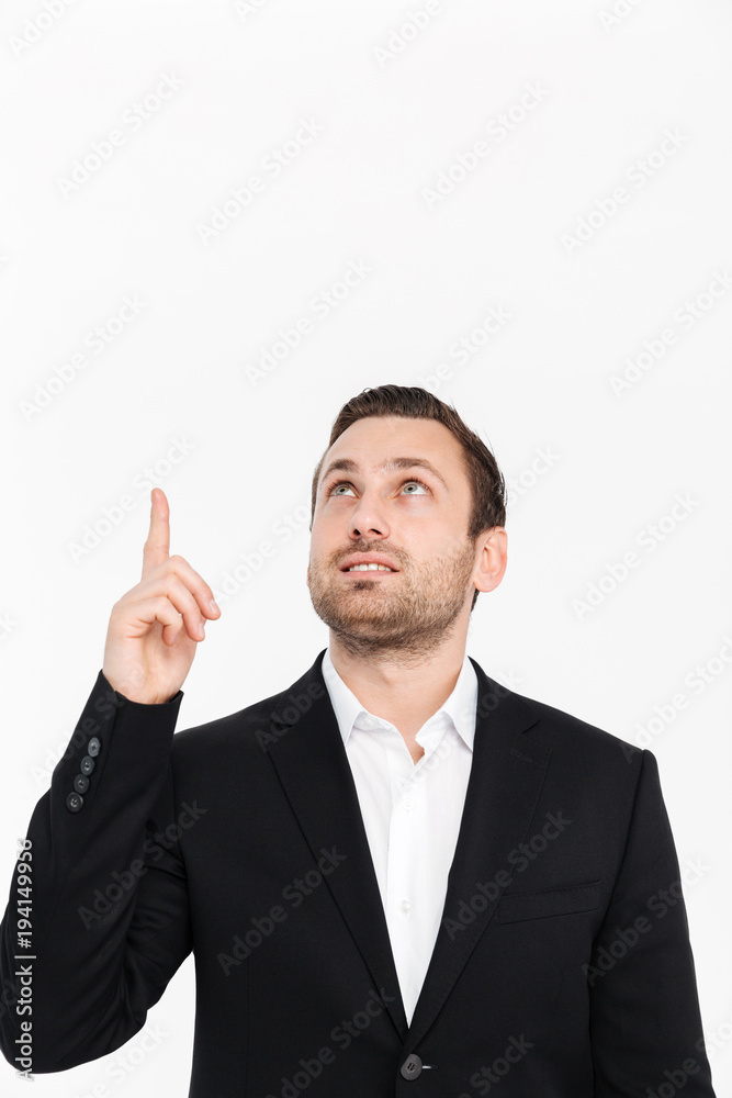 Portrait of smiling entrepreneur having stubble wearing black suit looking upward and pointing index finger up, isolated over white background copy space Portrait of smiling entrepreneur having stubble wearing black suit looking upward and pointing index finger up, isolated over white background copy space