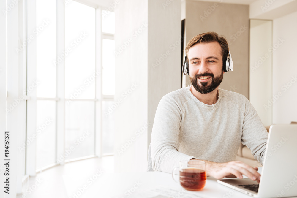 Photo of thoughtful man with short brown hair and beard, listening to music via wireless headphones and chatting on notebook Photo of thoughtful man with short brown hair and beard, listening to music via wireless headphones and chatting on notebook