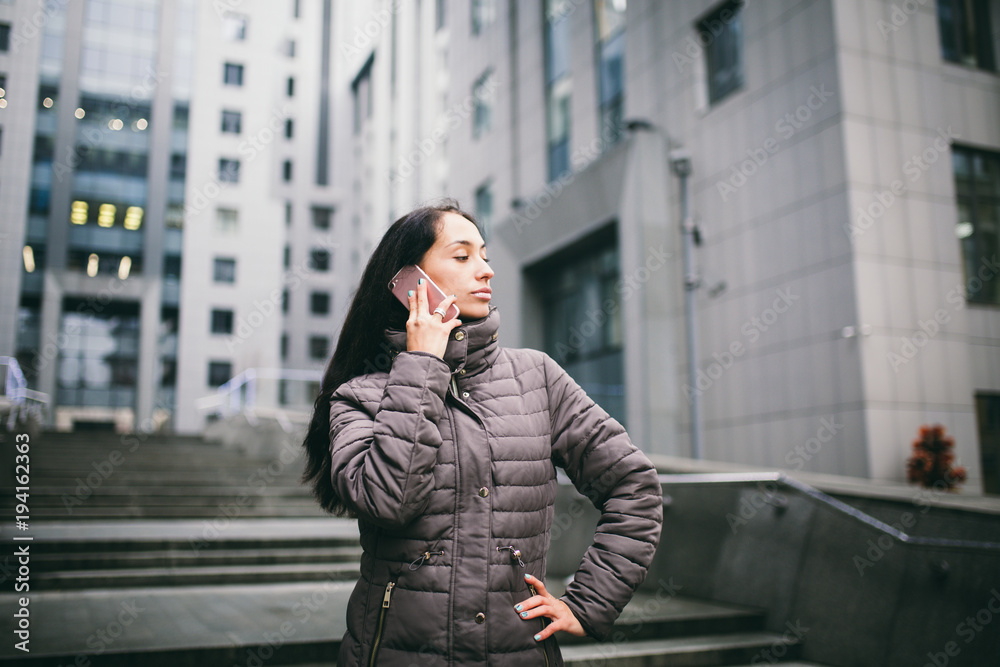 Fototapeta premium young girl talking on mobile phone in courtyard business center. girl with long dark hair dressed in winter jacket in cold weather speaks on phone on background buildings made of glass and concrete