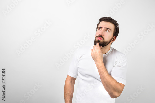 Young and hans dsome man is standing at the white wall and keeping his left hand close to the chin. He is thinking and looking to somewhere aside. Isolated on white background. photo