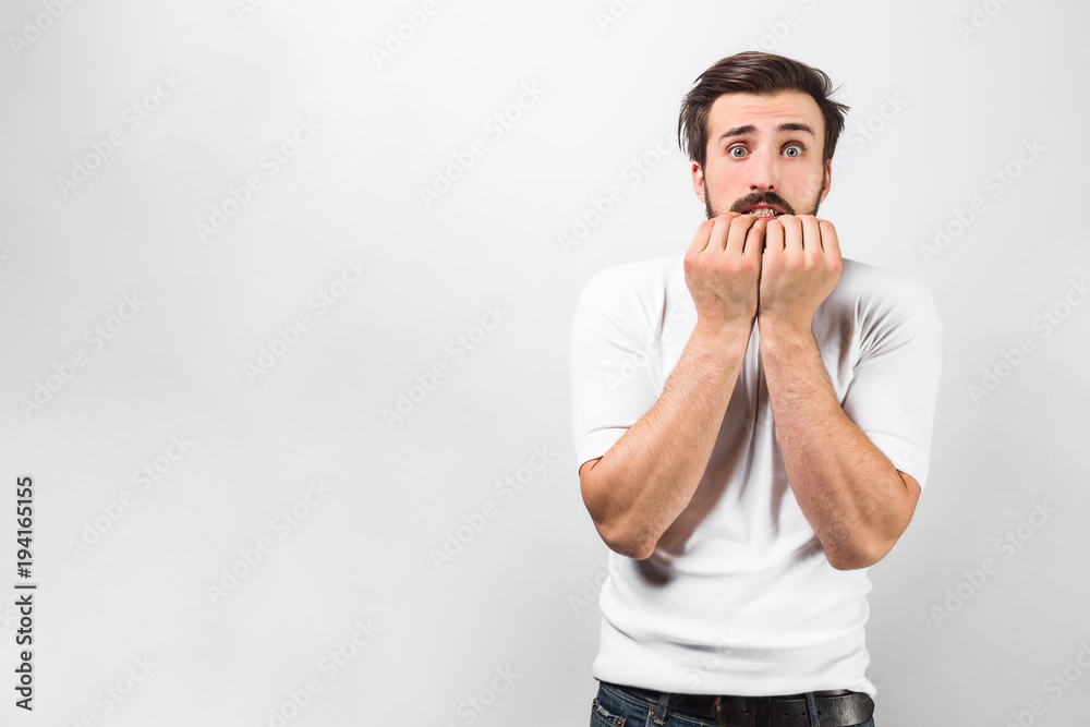 A picture of a guy standind near the white wall and holding his fingers close to the mouth. His sight is terrifying and full of horror. He is afraid of something. Isolated on white background. A picture of a guy standind near the white wall and holding his fingers close to the mouth. His sight is terrifying and full of horror. He is afraid of something. Isolated on white background.