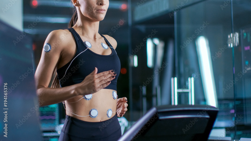 Beautiful Woman Athlete Runs on a Treadmill with Electrodes Attached to Her Body, Female Physician Uses Tablet Computer and Controls EKG Data Showing on Laboratory Monitors. Beautiful Woman Athlete Runs on a Treadmill with Electrodes Attached to Her Body, Female Physician Uses Tablet Computer and Controls EKG Data Showing on Laboratory Monitors.