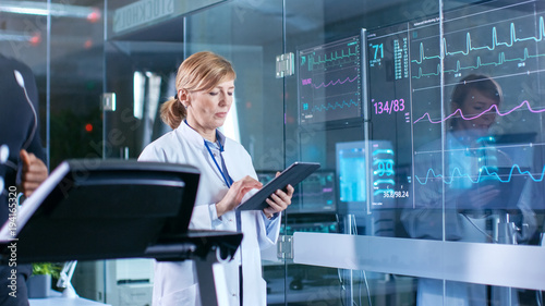 Male Athlete Runs on a Treadmill with Electrodes Attached to His Body while Sport Scientist Holds Tablet and Supervises EKG Status. In the Background Laboratory with High-Tech Equipment.