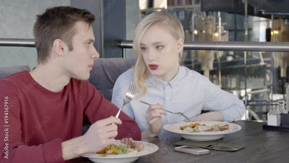Pair of colleagues are having lunch together in stylish restaurant, slow motion. Young man in bordo jumper and blond woman in blue shirt are sitting on velvet sofa at the wooden table, eating salad Pair of colleagues are having lunch together in stylish restaurant, slow motion. Young man in bordo jumper and blond woman in blue shirt are sitting on velvet sofa at the wooden table, eating salad