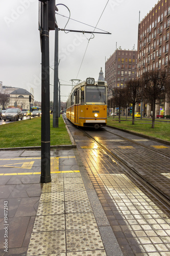 Budapest, Hungary - 02/22/2018: Old yellow tram, car traffic and buildings on city street. Transport and travel concept. Train rails and city road. Budapest landmark and transportation. Cityscape.