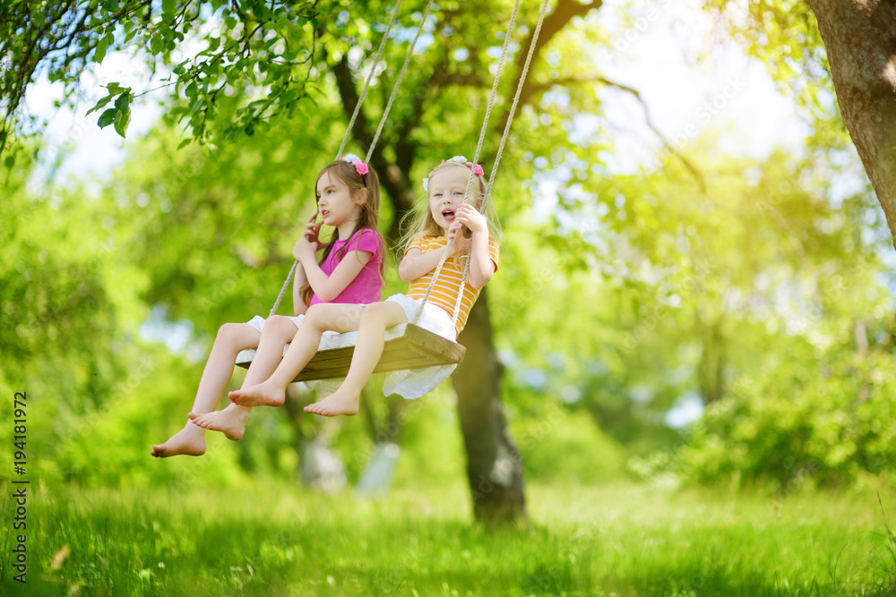 Two cute little sisters having fun on a swing together in beautiful summer garden on warm and sunny day outdoors Two cute little sisters having fun on a swing together in beautiful summer garden on warm and sunny day outdoors