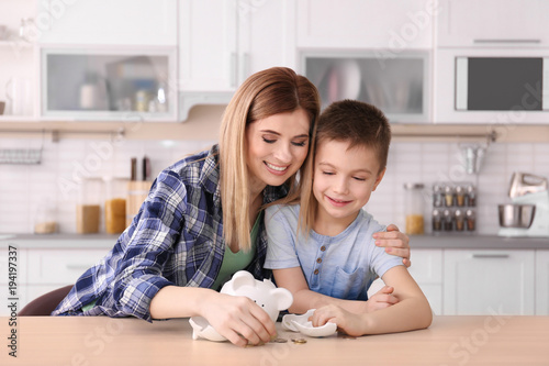 Mother and little son with broken piggy bank on table at home