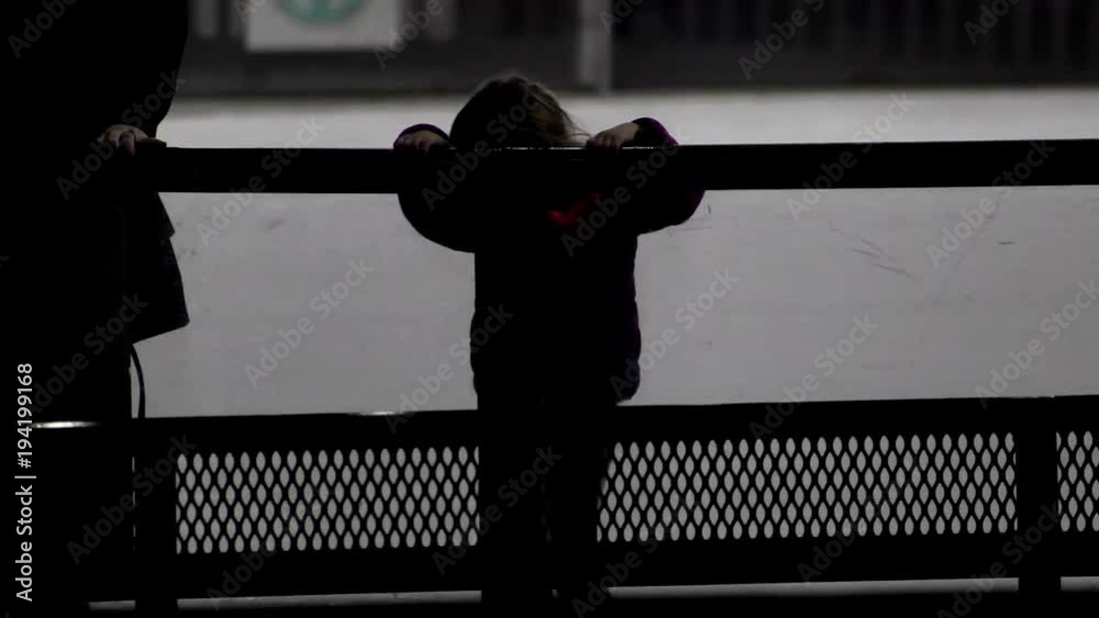 Little girl holds the railing of an outdoor ice skating rink, behind ...