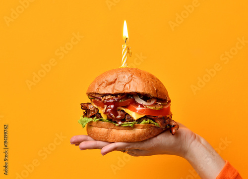 Obraz na płótnie Woman hands hold big burger barbeque sandwich with beef and lit candle for birthday party on yellow w salonie
