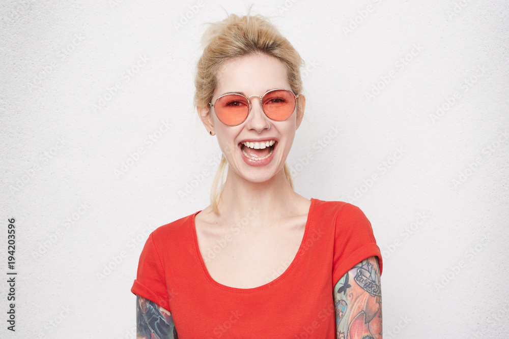 Portrait of pretty blonde girl laughs and looking directly into camera. Wears casual red t-shirt and red stylish glasses. Positive emotions. Pierced nose Portrait of pretty blonde girl laughs and looking directly into camera. Wears casual red t-shirt and red stylish glasses. Positive emotions. Pierced nose