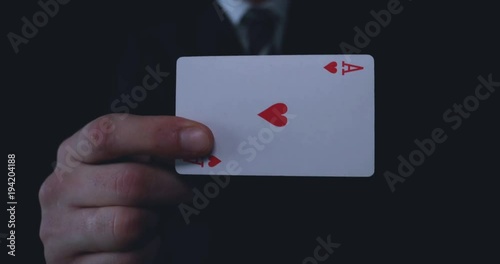 A businessman or magician holds a playing card (ace of hearts), in a suit, a black background.