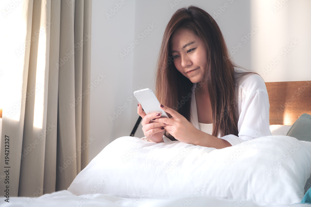 Portrait image of a beautiful Asian woman sitting on the bed , using and looking at mobile phone after woke up with feeling happy Portrait image of a beautiful Asian woman sitting on the bed , using and looking at mobile phone after woke up with feeling happy