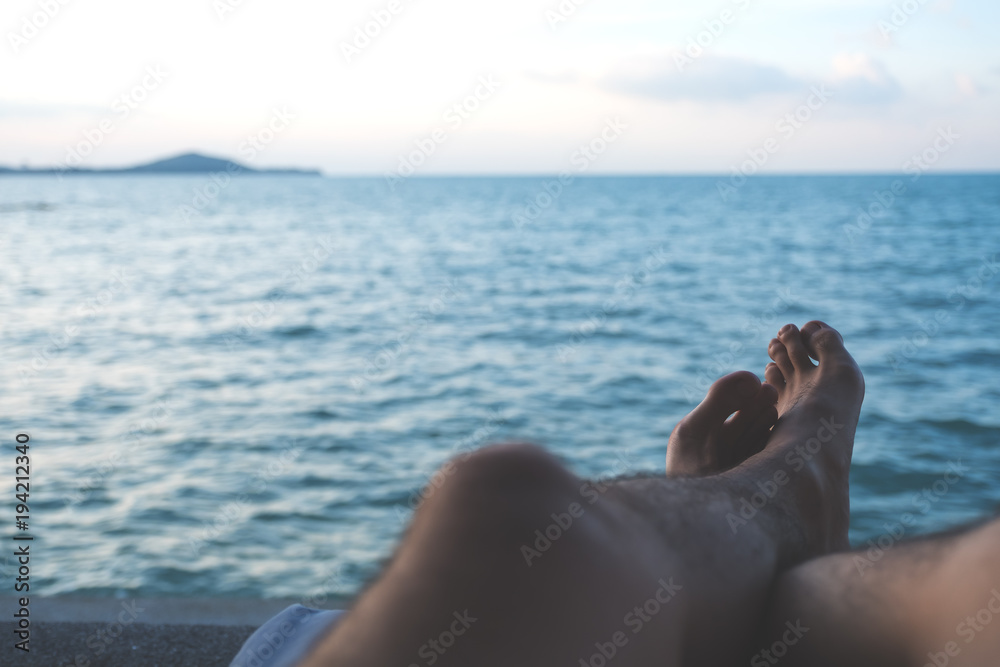 Closeup image of man's legs and foot while sitting by the sea with blue sky background
