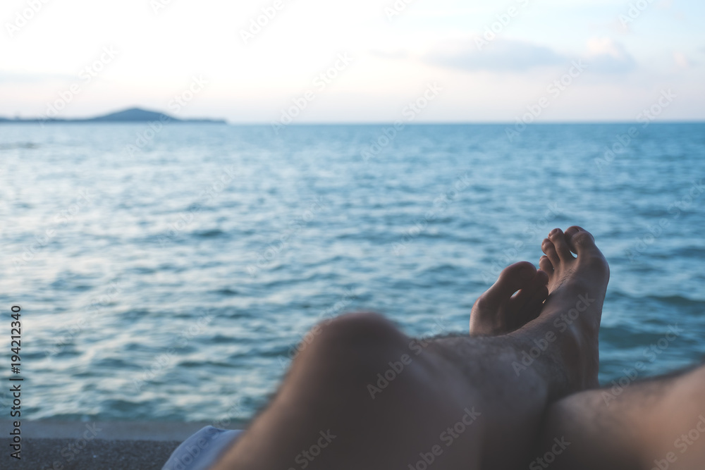 Fototapeta premium Closeup image of man's legs and foot while sitting by the sea with blue sky background