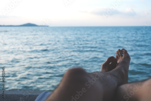 Closeup image of man's legs and foot while sitting by the sea with blue sky background