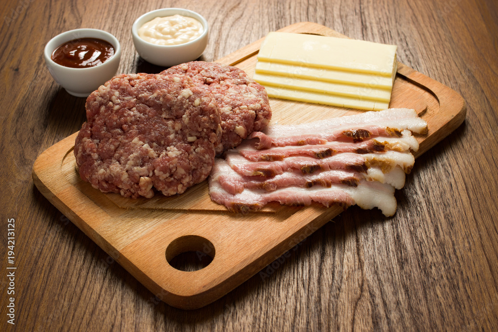 Ingredients for cooking burgers. Raw ground beef meat cutlets on wooden chopping board, cheese, bacon, over wooden background Ingredients for cooking burgers. Raw ground beef meat cutlets on wooden chopping board, cheese, bacon, over wooden background